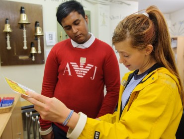 A young person in a yellow raincoat & long hair standing next to a young person wearing a red jumper. They are concentrating on a document, a map, an artwork, or instructions.