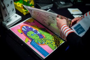 Hands leafing through a brightly coloured book in which a cartoon green man is depicted in a pink room.