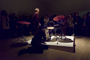 'The Legal Team, The Singer, The Chorus and The Gait', Paul Abbott and Cara Tolmie, Performance, 2014. The spot-lit artist wearing a red velvet dress and a long plait performs on a stage with a drum kit. There audience are in the dark.