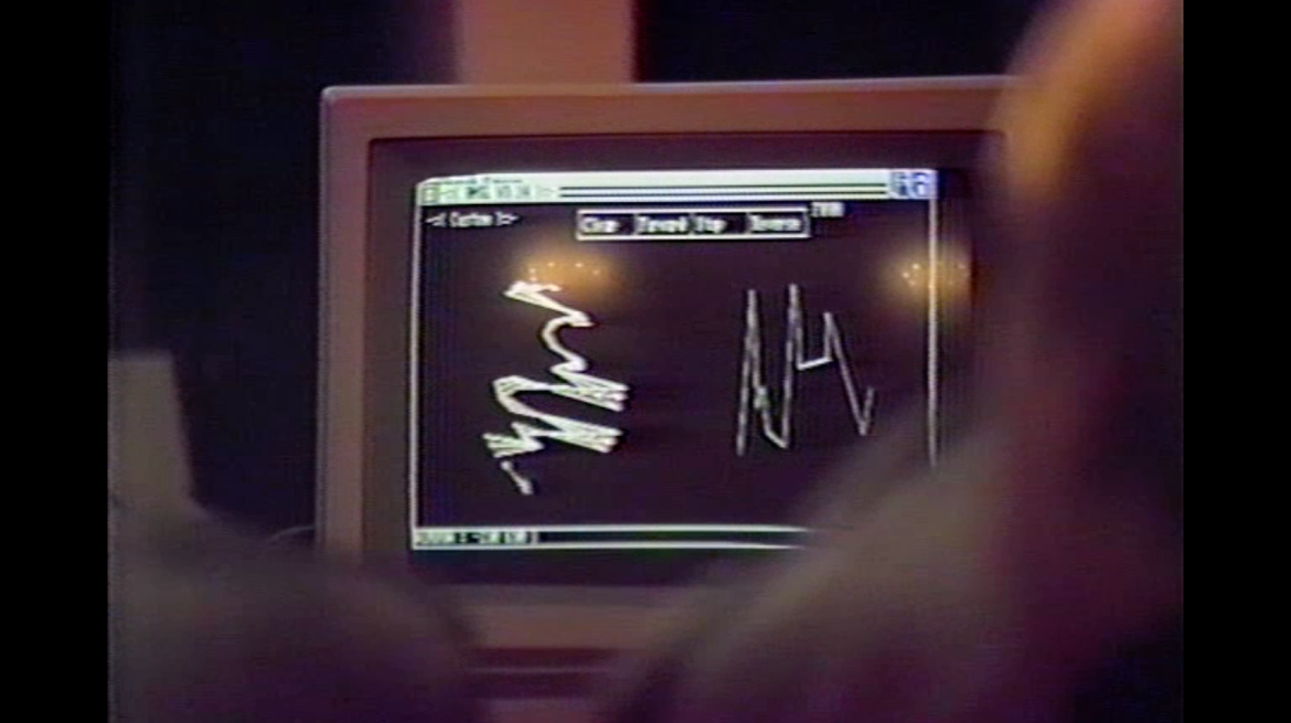 Grainy film still featuring two out of focus figures looking at a computer screen displaying indecipherable charts and lines.