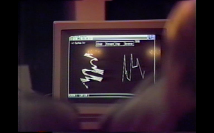 Grainy film still featuring two out of focus figures looking at a computer screen displaying indecipherable charts and lines.