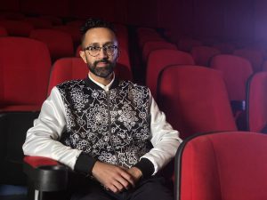 Image of Hetain Patel sitting in a red theatre seat. He is wearing black glasses and a black and white patterned jacket.