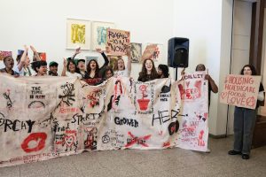 A group of young adults holding a banner with spray painted images and words such as 'growth' and 'anarchy starts here'. The young people punch the air and shout.