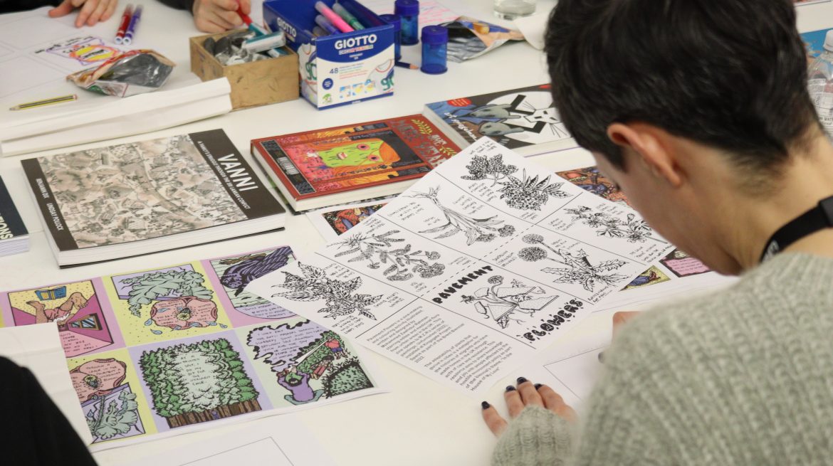 A person sat a table covered in unfolded zine designs. They are drawing.