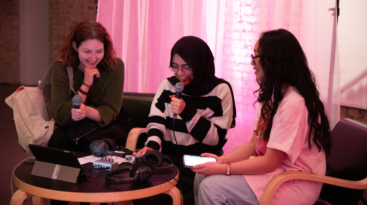 3 young people sit together and soeak into a microphone in front of a pink curtain