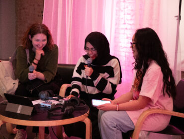 3 young people sit together and soeak into a microphone in front of a pink curtain